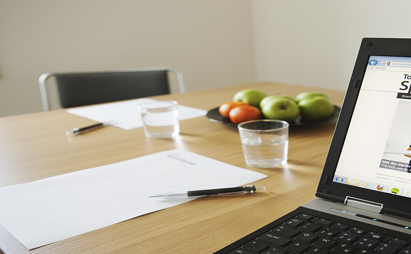 Bilden visar ett bord i ett konferensrum. Till höger på bordet står en dator och till vänster ligger ett vitt papper och en penna. I bakgrunden står ett fruktfat och två glas vatten. 