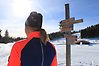 Bild på en skidåkare som står i solen på skidstation. Till höger i bild ser man en trästolpe med skyltar på som visar våra åt vilka håll våra motionsspår går. 